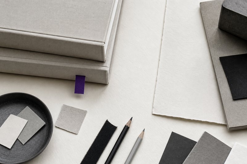 The corner of a reading-room desk seen from above with a squared stack of large reference volumes, their covers blank, a violet bookmark protruding.